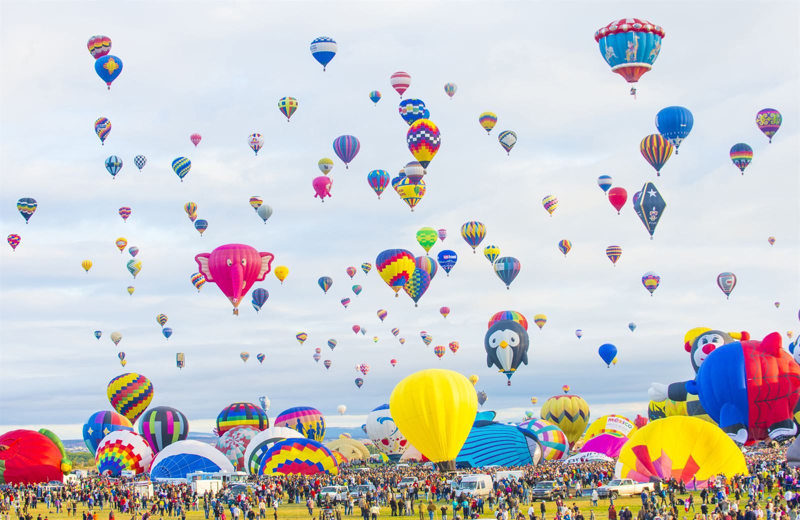 Hundreds of hot-air balloons, including special-shape balloons, rising over a crowded launch field in Albuquerque