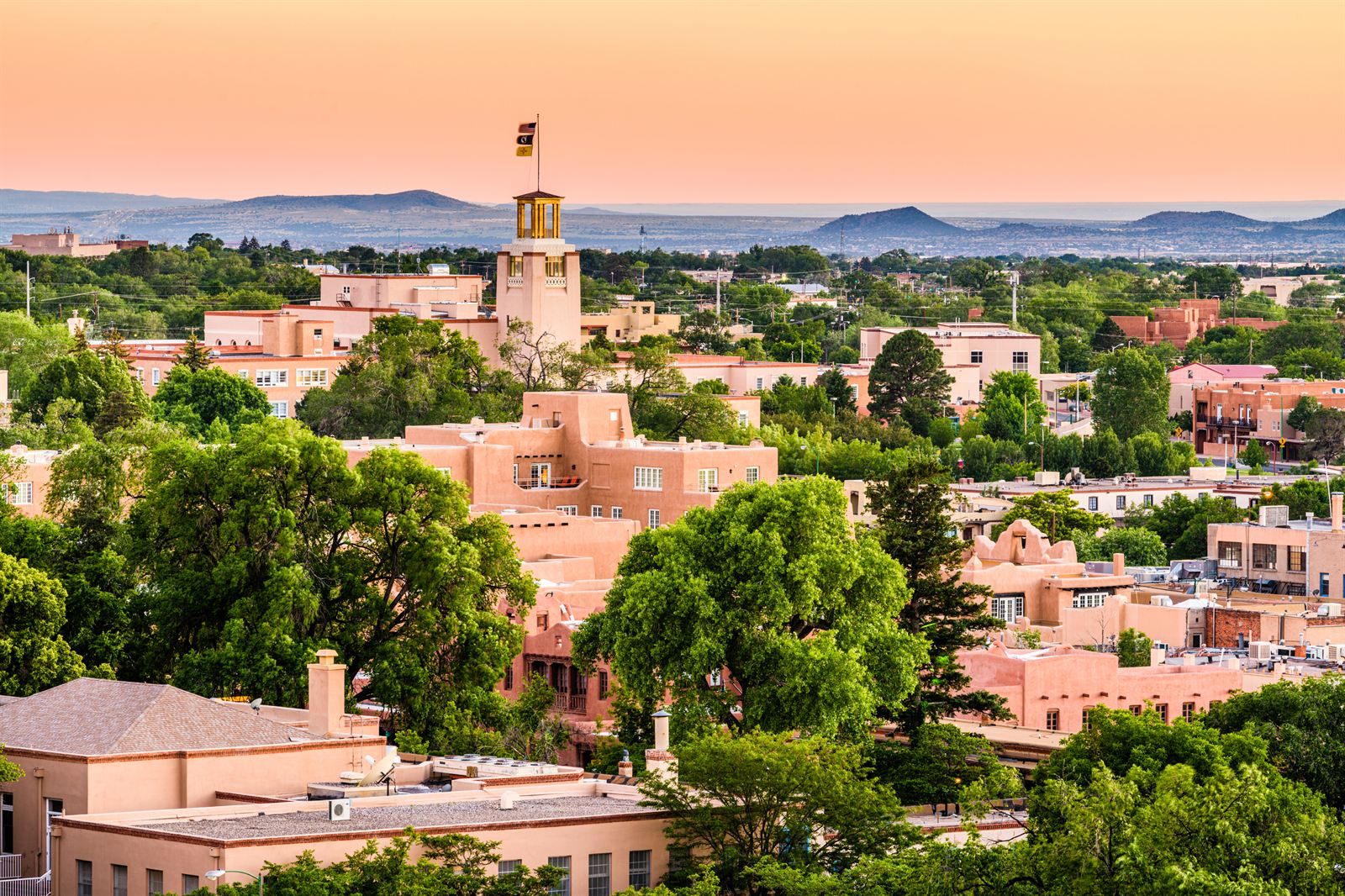 Low adobe-style buildings and green trees across Santa Fe at sunset, with mountains on the horizon