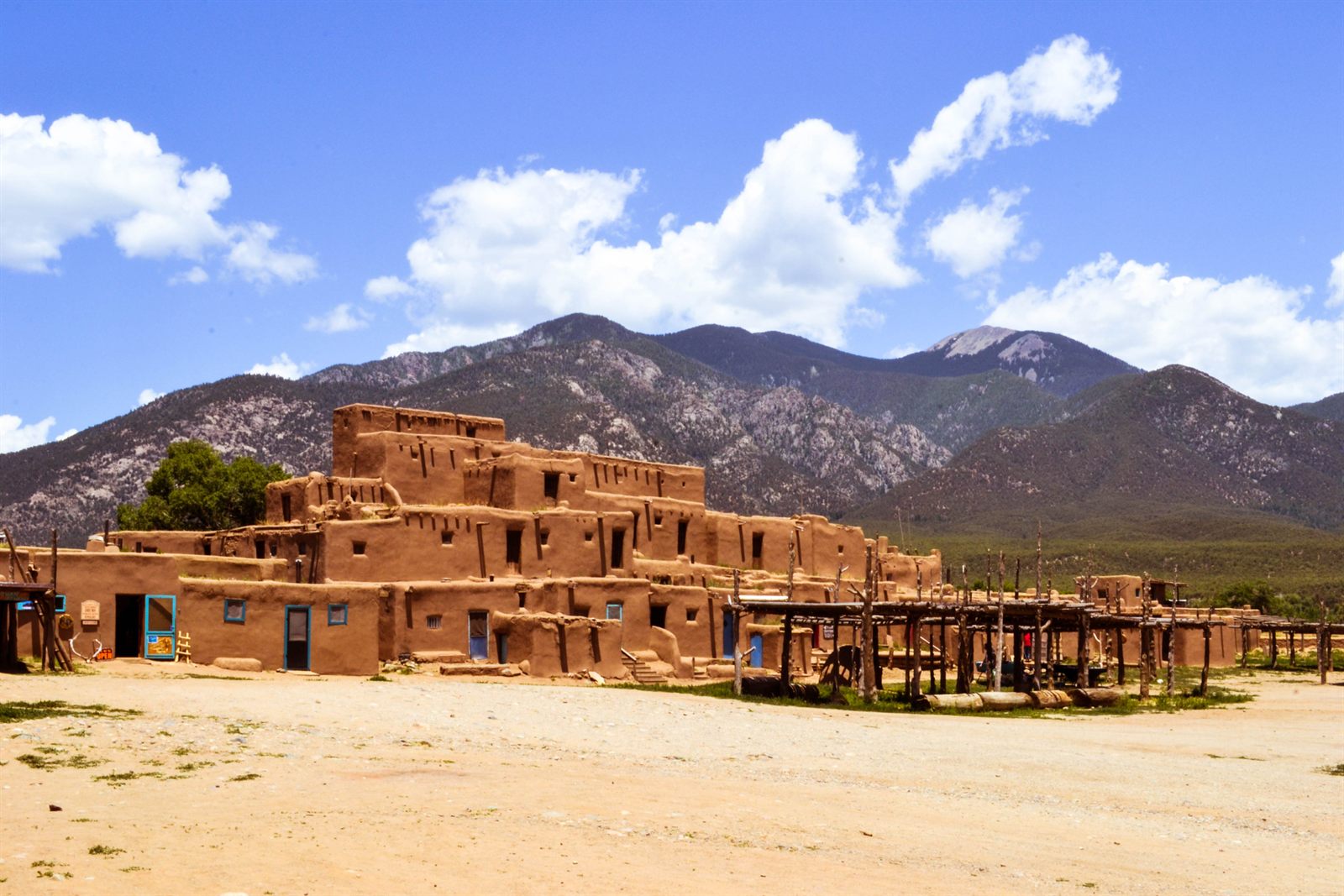 The multistory adobe homes of Taos Pueblo under a blue sky, with mountains behind